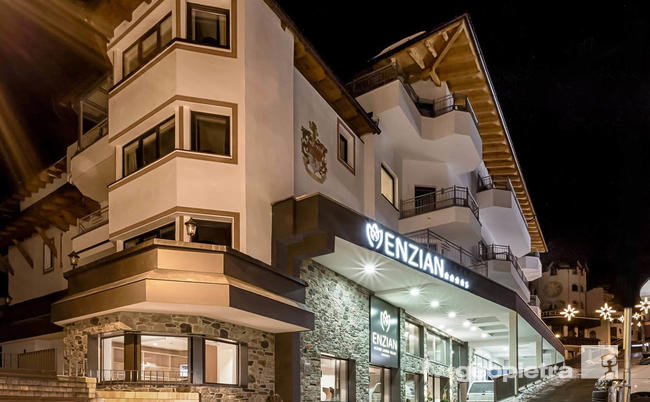 Luxury multi-story building characterized by a luminous sign with the word "ENZIAN" and five stars next to it, suggesting a five-star hotel. The lower part of the building features Geopietra reconstructed stone cladding. The building is surrounded by other structures and is photographed at night, illuminated by artificial lights.